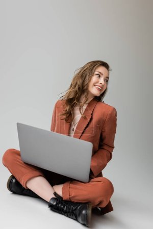 cheerful young woman in golden necklace sitting with crossed legs in  trendy suit, using laptop, looking away while working remotely on grey background, freelancer, digital nomad 