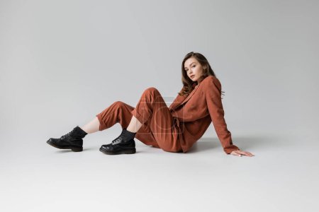 Photo for Full length of trendy model with brunette and wavy hair sitting in trendy and oversize suit with terracotta blazer, pants and black boots, looking at camera on grey background, stylish young woman - Royalty Free Image