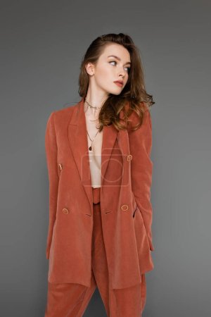 young shirtless woman with brunette and wavy hair posing in trendy and terracotta suit with oversize blazer and golden necklace, looking away on grey background,  stylish pose, sexy model 
