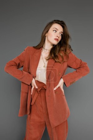 young shirtless woman with brunette and wavy hair posing with hands on hips in trendy and oversize suit with blazer and golden necklace, looking away on grey background,  stylish pose, sexy model 