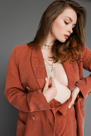 Photo for Sexy young woman with brunette and wavy hair posing in trendy and oversize suit with terracotta blazer and golden necklace, looking away on grey background, stylish pose, hand on hip - Royalty Free Image
