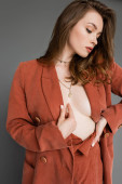 sexy young woman with brunette and wavy hair posing in trendy and oversize suit with terracotta blazer and golden necklace, looking away on grey background, stylish pose, hand on hip  puzzle #656946560