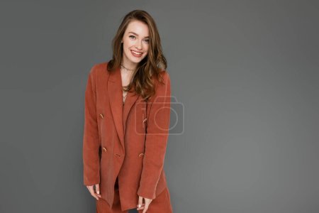Photo for Happy young woman with brunette and wavy hair posing in trendy and oversize suit with terracotta blazer and smiling while looking at camera on grey background, stylish pose, trendy chic - Royalty Free Image