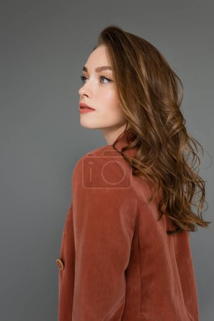 Porträt einer anspruchsvollen jungen Frau mit brünetten Haaren, die im braunen und trendigen Anzug mit Blazer posiert, während sie vor grauem Hintergrund im Studio wegschaut, hübsches Modell, Locken, trendige Frisur 