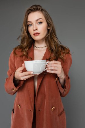 kultivierte junge Frau mit brünetten Haaren trägt braunen und trendigen Anzug mit Blazer und hält eine Tasse Kaffee in der Hand, während sie vor grauem Hintergrund in die Kamera blickt, Work-Life-Balance 