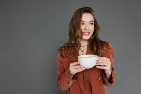 happy young woman with brunette hair wearing brown and trendy suit with blazer and holding cup of coffee while looking away on grey background, work-life balance