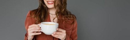 vue recadrée d'une jeune femme heureuse aux cheveux bruns portant un costume marron et tendance avec un blazer et tenant une tasse de café sur fond gris, équilibre travail-vie privée, bannière 
