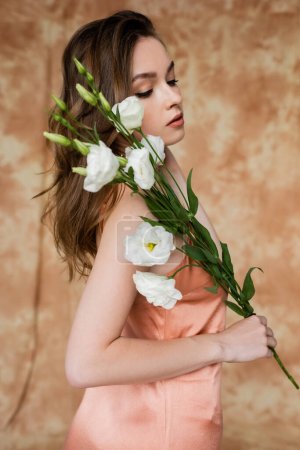 portrait de brune et jeune femme en soie rose slip robe tenant des fleurs d'eustomie tout en se tenant debout sur fond beige marbré, sensualité, élégance, sophistication 
