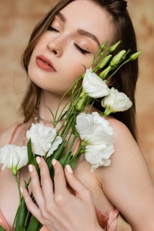 portrait de femme rêveuse, brune et jeune aux yeux fermés tenant des fleurs d'eustomie debout et posant sur fond beige marbré, sensualité, élégance, sophistication 