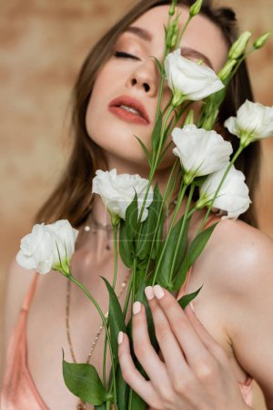 portrait of sensitive and young woman in pink silk slip dress holding eustoma flowers while standing with closed eyes on mottled beige background, sensuality, elegance, sophistication 