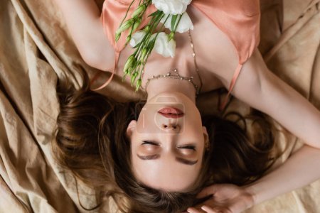 top view of alluring young woman in pink silk slip dress lying with closed eyes on linen fabric with delicate white flowers on beige background, sensuality, upside down view 