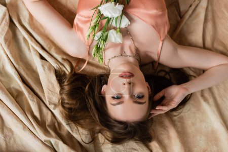 Foto de Vista superior de la atractiva mujer joven en vestido de seda rosa deslizamiento acostado en tela de lino con delicadas flores blancas y mirando a la cámara sobre fondo beige, sensualidad, vista al revés - Imagen libre de derechos