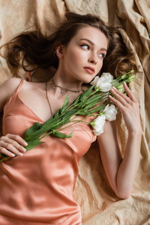 top view of attractive young woman in pink silk slip dress lying on linen fabric and holding delicate white flowers on beige background, sensuality, elegance, eustoma, looking away 