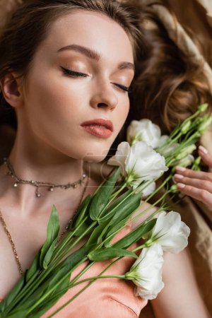 top view of attractive young woman in pink silk slip dress lying with closed eyes on linen fabric and holding white flowers on beige background, sensuality, elegance, eustoma, portrait 