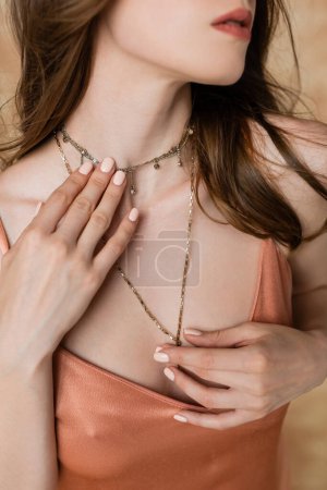 Photo for Cropped view of captivating young woman with brunette hair touching neck with sophisticated golden necklace while posing in pink slip dress on mottled beige background, sensuality - Royalty Free Image