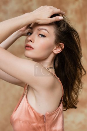 sexy mujer joven con el pelo morena posando en vestido de deslizamiento rosa con las manos cerca del pelo sobre fondo beige moteado, sofisticación, sensualidad, elegancia, mirando a la cámara  
