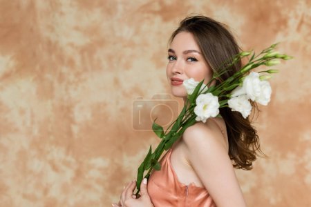 glückliche junge Frau mit brünetten Haaren posiert in rosa und seidenem Slip-Kleid und hält weiße Eustoma-Blumen auf fleckigem beigem Hintergrund, Sinnlichkeit, Raffinesse, Eleganz, blickt in die Kamera