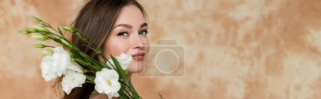 portrait de jeune femme heureuse aux cheveux bruns souriant tout en tenant des fleurs d'eustomie blanches sur fond beige marbré, sensualité, sophistication, élégance, regardant la caméra, bannière 