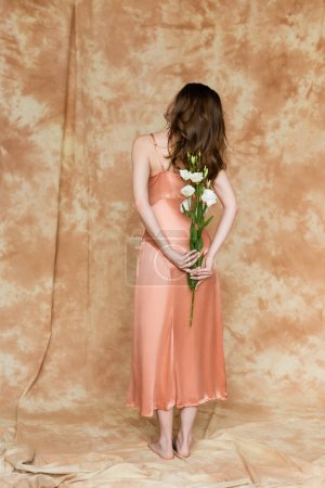 back view of graceful young woman with brunette hair standing in slip dress on linen fabric and holding white flowers behind back on mottled beige background, sensuality, elegance, full length