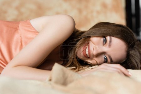 Photo for Positive and graceful young woman with brunette hair lying in silk slip dress and looking at camera on blurred and linen fabric on mottled beige background, sophistication, sensuality, elegance - Royalty Free Image