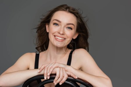 portrait of beautiful and happy young woman with brunette hair and blue eyes smiling while leaning on chair back and looking at camera isolated on grey background, sensuality, grace, feminine beauty 
