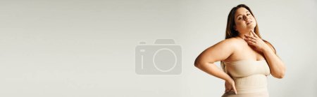 Femme avec un corps de taille plus touchant son cou et regardant la caméra tout en posant avec la main sur la hanche en haut bustier beige et sous-vêtements en studio isolé sur fond gris, corps positif, bannière 