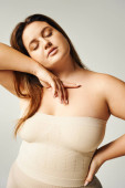 sensual woman with plus size body touching her cheek with hand while posing with closed eyes in beige strapless top in studio isolated on grey background, body positive, self-love  t-shirt #656983510