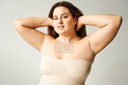 sensual woman with plus size body touching hair and posing in beige strapless top in studio isolated on grey background, body positive, self-love, looking at camera, self-esteem 