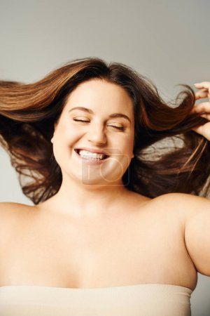 Porträt einer strahlenden Frau mit Plus-Size-Körper und geschlossenen Augen, die Haare berührt und mit nackten Schultern auf grauem Hintergrund im Studio posiert, körperbetont, selbstverliebt 