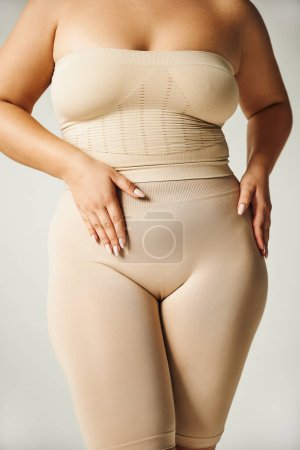 Ausgeschnittene Ansicht von unkenntlich Plus Size Frau posiert in trägerlosem Top mit nackten Schultern und Unterwäsche isoliert auf grauem Hintergrund im Studio, Körper positiv, Selbstliebe, Hand auf Bauch 