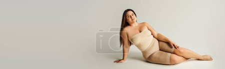 Barfüßige und selbstbewusste Frau in trägerlosem Top mit nackten Schultern und Unterwäsche, die im Studio auf grauem Hintergrund posiert, körperbetont, selbstverliebt, plus Größe, Banner 