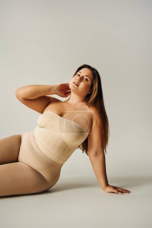 Selbstbewusste Frau in trägerlosem Top mit nackten Schultern und Unterwäsche posiert im Studio auf grauem Hintergrund, körperbetont, selbstverliebt, plus Größe, Figurentyp 