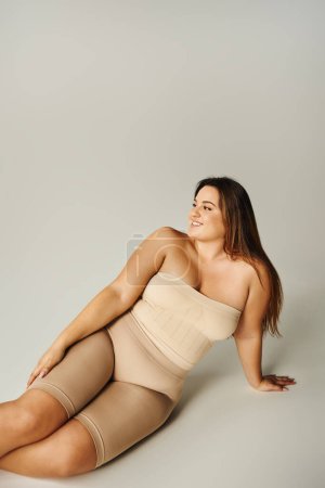 cheerful woman in strapless top with bare shoulders and underwear posing while sitting in studio on grey background, body positive, self-love, plus size, figure type 