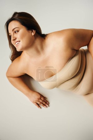 Draufsicht einer lächelnden Frau in trägerlosem Top mit nackten Schultern und Unterwäsche, die im Studio auf grauem Hintergrund posiert, körperbetont, selbstverliebt, plus Größe, Figurentyp, wegschauen 