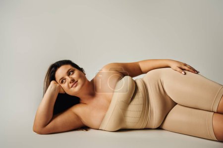 brünette Frau mit Plus-Size-Körper in trägerlosem Top mit nackten Schultern und Unterwäsche posiert im Studio auf grauem Hintergrund, körperbetont, selbstverliebt, entspannend, wegschauend 