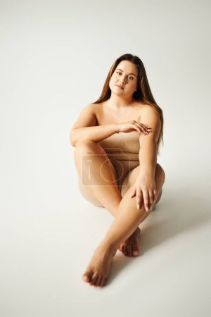 Barfuß-Model mit Plus-Size-Körper in trägerlosem Top mit nackten Schultern und Unterwäsche posiert im Studio auf grauem Hintergrund, körperbetont, selbstverliebt, entspannend, in die Kamera schauend 
