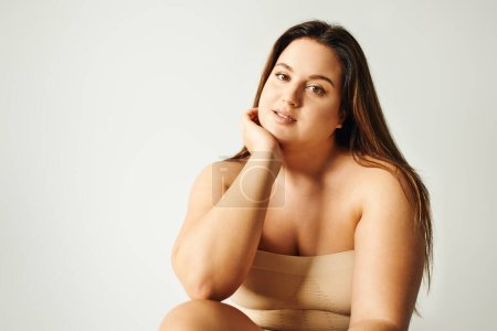 brunette model with plus size body in strapless top with bare shoulders posing while sitting in studio on grey background, body positive, self-love, relaxing, looking at camera 