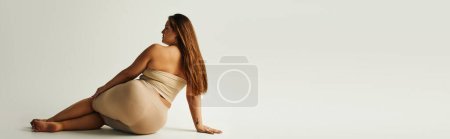back view of barefoot woman with plus size body in strapless top with bare shoulders posing while sitting in studio on grey background, body positive, tattoo translation: harmony, banner 