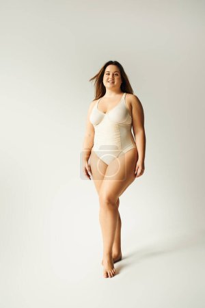 full length of barefoot and plus size woman in beige bodysuit posing while standing in studio on grey background, body positive, figure type, self-esteem, smiling while looking at camera 