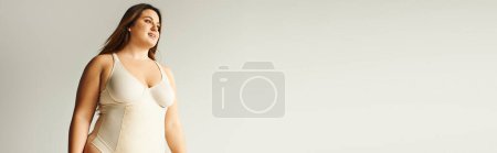 happy plus size woman with natural makeup posing in beige bodysuit while standing in studio on grey background, body positive, figure type, self-esteem, smiling while looking away, banner 