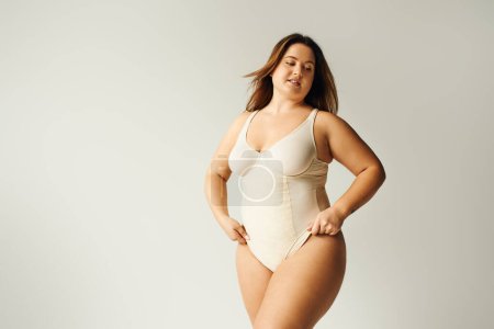 tattooed and brunette curvy woman with plus size body posing in beige bodysuit while standing in studio on grey background, body positive, figure type, self-esteem, tattoo translation: harmony 