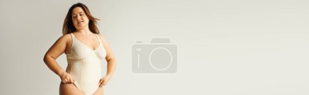 Téléchargez les photos : Femme courbée tatouée et brune avec corps de taille plus posant en body beige, debout en studio sur fond gris, corps positif, type de figure, estime de soi, traduction de tatouage : harmonie, bannière - en image libre de droit