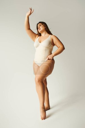 full length of brunette curvy woman with plus size body posing in beige bodysuit while standing with raised hand in studio on grey background, body positive, figure type, smiling while looking away 