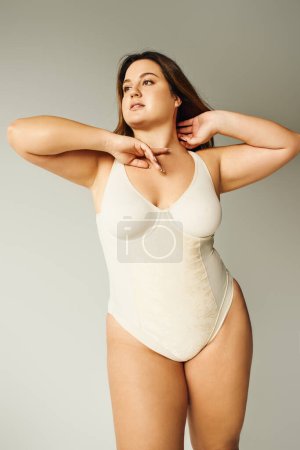 brunette curvy woman with plus size body posing in beige bodysuit while standing and touching hair in studio on grey background, body positive, figure type, looking away 