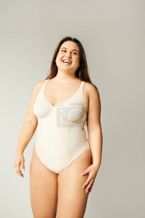 portrait of positive and curvy woman with plus size body posing in beige bodysuit while laughing on grey background, body positive, figure type, looking away while standing in studio  mug #656984280