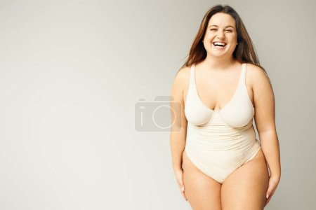 Porträt einer glücklichen und kurvigen Frau mit Plus-Size-Körper, die im beigen Body posiert, während sie auf grauem Hintergrund lacht, körperbetont, figurbetont, im Studio in die Kamera blickt 