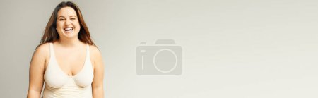Téléchargez les photos : Portrait de femme heureuse et courbée avec un corps de taille plus posant en body beige tout en riant sur fond gris, corps positif, type de figure, regardant la caméra debout en studio, bannière - en image libre de droit