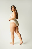 full length of brunette and curvy woman wearing beige bodysuit and standing with bare feet on grey background, self-esteem, figure type, looking at camera, body positivity movement  Poster #656984332