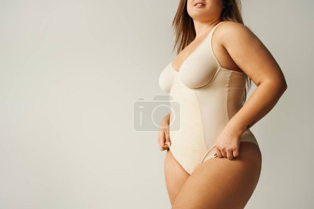partial view of curvy woman wearing beige bodysuit and standing with hand on hip isolated on grey background, self-confidence, figure type, body positivity movement, tattoo translation: harmony 