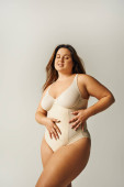 brunette and curvy woman with tattoo wearing beige bodysuit and standing with hands on waist on grey background, body positive, figure type, body positivity movement, closed eyes  Poster #656984494